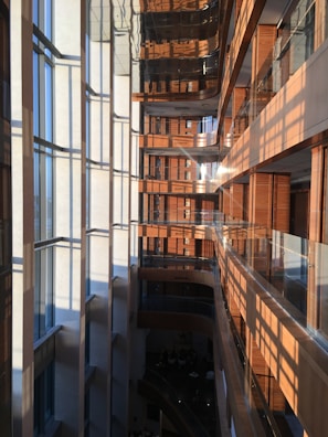 A modern multi-story building interior with a large vertical atrium. Glass windows on the left side allow sunlight to stream in, creating a pattern of light and shadows on the opposite wooden walls and balconies. The building features sleek surfaces, clean lines, and a mix of natural wood and glass materials.