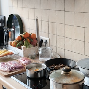 A kitchen countertop with various cooking ingredients and utensils. Raw cuts of meat are placed on a wooden board next to a pot cooking an egg. A frying pan contains mushrooms, and there is a glass jar of salt nearby. A bowl of fresh vegetables, including carrots and persimmons, is visible. The backsplash features beige tiles, and there is a colander and other kitchen items on the counter.