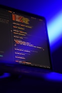 A computer screen displays lines of colorful code in a dimly lit room. The screen is angled to show the glowing text against a dark background, with blue ambient lighting creating a tech-focused atmosphere.