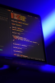 A computer screen displays lines of colorful code in a dimly lit room. The screen is angled to show the glowing text against a dark background, with blue ambient lighting creating a tech-focused atmosphere.