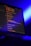 A computer screen displays lines of colorful code in a dimly lit room. The screen is angled to show the glowing text against a dark background, with blue ambient lighting creating a tech-focused atmosphere.