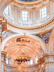 The interior features an ornate and intricately decorated ceiling with detailed gold embellishments and fresco paintings. Large windows allow natural light to illuminate the space, highlighting the elaborate architectural details and artwork. The central part of the ceiling is adorned with a grand chandelier, adding to the opulence of the interior design.