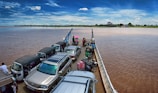 A ferry is carrying multiple vehicles and people across a wide river under a clear blue sky. The vehicles include cars, tuk-tuks, and motorcycles, and people are seen standing along the sides of the ferry. The background features a distant shoreline with green vegetation and buildings.