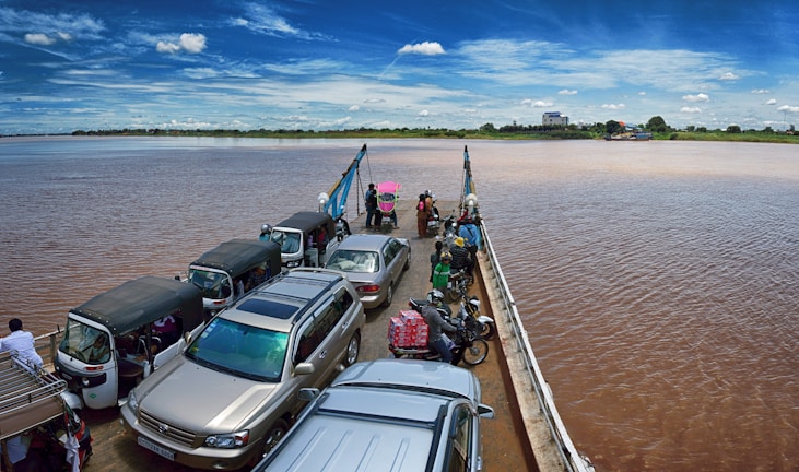A ferry is carrying multiple vehicles and people across a wide river under a clear blue sky. The vehicles include cars, tuk-tuks, and motorcycles, and people are seen standing along the sides of the ferry. The background features a distant shoreline with green vegetation and buildings.