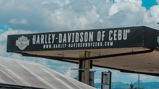 A sign for Harley-Davidson of Cebu is prominently displayed against a background of a partly cloudy sky. The sign is black with white text and features the Harley-Davidson logo. Below the main text, there is a web address also in white letters.