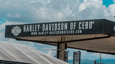 A sign for Harley-Davidson of Cebu is prominently displayed against a background of a partly cloudy sky. The sign is black with white text and features the Harley-Davidson logo. Below the main text, there is a web address also in white letters.