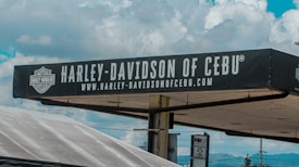 A sign for Harley-Davidson of Cebu is prominently displayed against a background of a partly cloudy sky. The sign is black with white text and features the Harley-Davidson logo. Below the main text, there is a web address also in white letters.