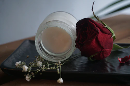 Minimalist black vase holding a single red rose on a bone-colored surface beside a delicate jar of moisturizer.