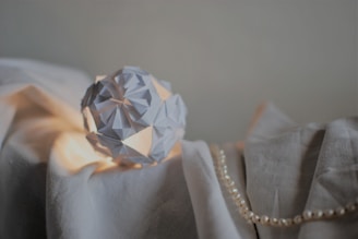 An intricately folded paper origami sphere is placed on a gently draped white fabric. The scene is softly illuminated, accentuating the shadows and textures. A string of pearls lays beside the paper sculpture, adding a touch of elegance.