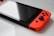 A close-up view of a handheld gaming console with a rectangular screen and bright red controllers on the sides. The buttons and joysticks are visible, and the surface of the console is glossy, reflecting light.