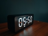 A digital clock with a sleek black frame showing time and date on a kitchen counter.