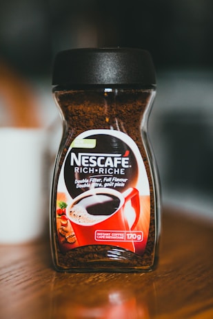A jar of Nescafe instant coffee is placed on a wooden surface. The jar features the label Nescafe Rich in English and French, with details about its rich, double-filtered flavor. A graphic of a red coffee mug filled with coffee is prominently displayed on the front.