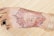 A hand adorned with intricately designed henna, featuring floral patterns and crescent motifs. The design is detailed and symmetrical, blending traditional elements with text elements in the center. The fingers also have smaller, simpler designs.