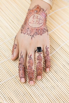 A henna design intricately applied on the back of a hand featuring floral patterns and calligraphy. The fingers are adorned with detailed henna art, and a black ring is worn on one of the fingers. The background consists of a textured bamboo mat.
