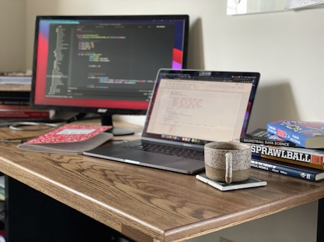 A workspace featuring a wooden desk with a laptop displaying code, a monitor showing programming work, several books stacked to the right, and a speckled mug on a coaster. The environment conveys a tidy and focused setup suitable for programming tasks.