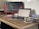 A workspace featuring a wooden desk with a laptop displaying code, a monitor showing programming work, several books stacked to the right, and a speckled mug on a coaster. The environment conveys a tidy and focused setup suitable for programming tasks.