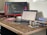A workspace featuring a wooden desk with a laptop displaying code, a monitor showing programming work, several books stacked to the right, and a speckled mug on a coaster. The environment conveys a tidy and focused setup suitable for programming tasks.