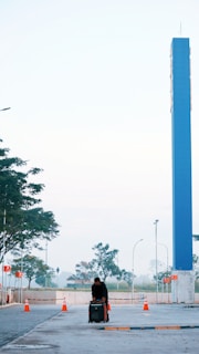 A person wearing dark clothing leans over a piece of luggage on a wide, paved area. Several bright orange traffic cones are arranged along the sides. A tall blue pillar stands prominently in the background. The scene is surrounded by trees and streetlights under a clear sky.