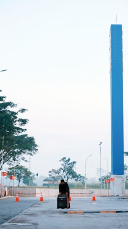 A person wearing dark clothing leans over a piece of luggage on a wide, paved area. Several bright orange traffic cones are arranged along the sides. A tall blue pillar stands prominently in the background. The scene is surrounded by trees and streetlights under a clear sky.