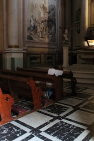 A person is kneeling in prayer at a wooden pew inside a church. The church interior features ornate architecture with tall columns and a large painting depicting a religious scene on the wall. There is a statue of an angel nearby, and sunlight is streaming through, illuminating parts of the floor and pew.