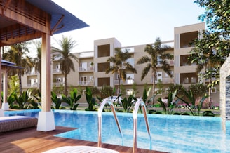 A luxurious residential complex is visible with a modern swimming pool in the foreground. Palm trees and a variety of lush green plants decorate the poolside area, while lounge chairs are placed under shaded wooden structures. The multi-story buildings in the background have balconies and large windows, giving the place a resort-like feel.