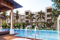 A luxurious residential complex is visible with a modern swimming pool in the foreground. Palm trees and a variety of lush green plants decorate the poolside area, while lounge chairs are placed under shaded wooden structures. The multi-story buildings in the background have balconies and large windows, giving the place a resort-like feel.