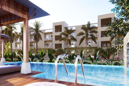 A luxurious residential complex is visible with a modern swimming pool in the foreground. Palm trees and a variety of lush green plants decorate the poolside area, while lounge chairs are placed under shaded wooden structures. The multi-story buildings in the background have balconies and large windows, giving the place a resort-like feel.