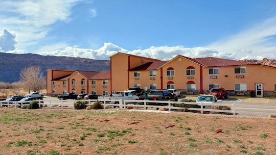 A two-story hotel building with a warm, orange exterior and red roofing. The structure has several windows and is surrounded by parked cars in a lot. A white fence encloses part of the area, with sparse greenery and rocky terrain in the foreground. A backdrop of rocky mountains and a partly cloudy sky can be seen in the distance.