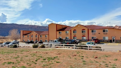 A two-story hotel building with a warm, orange exterior and red roofing. The structure has several windows and is surrounded by parked cars in a lot. A white fence encloses part of the area, with sparse greenery and rocky terrain in the foreground. A backdrop of rocky mountains and a partly cloudy sky can be seen in the distance.
