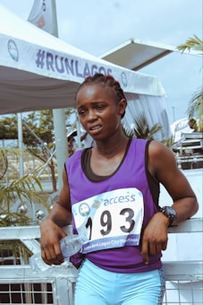 An athlete wearing a purple sports jersey with the number 193 stands outdoors, holding a water bottle. The backdrop includes a tent with the hashtag #RUNLAGOS, indicating a marathon event. Palm trees and a metal railing are visible nearby.