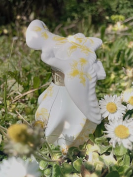 A porcelain figurine designed with yellow floral patterns is placed among green foliage and white daisies. The figurine features intricate details and has a soft sheen catching the sunlight.