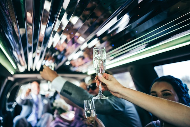Inside a limousine, a group of people raising champagne glasses in a toast. The interior features reflective surfaces and green ambient lighting. The atmosphere suggests a celebratory occasion.