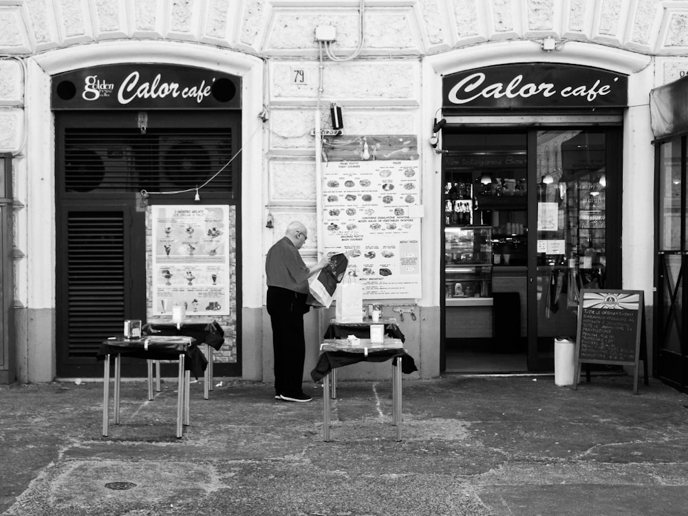 grayscale photo of man in black t-shirt and black pants standing near store