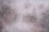 Rough, textured concrete wall with patches of discoloration and faint marks. The surface has a weathered appearance with various shades of gray and subtle hints of brown.