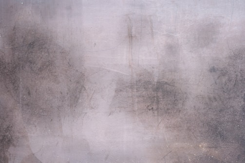 Rough, textured concrete wall with patches of discoloration and faint marks. The surface has a weathered appearance with various shades of gray and subtle hints of brown.