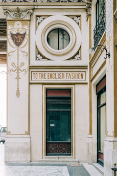An ornate architectural facade featuring elegant designs and gold accents. A prominent circular window and a sign reading 'To the English Fashion' are focal points. Intricate detailing and embellishments are present, contributing to an elegant and vintage aesthetic.
