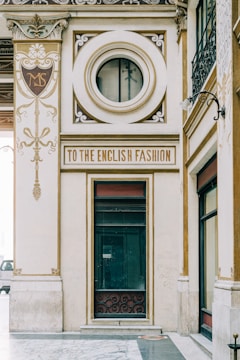 An ornate architectural facade featuring elegant designs and gold accents. A prominent circular window and a sign reading 'To the English Fashion' are focal points. Intricate detailing and embellishments are present, contributing to an elegant and vintage aesthetic.