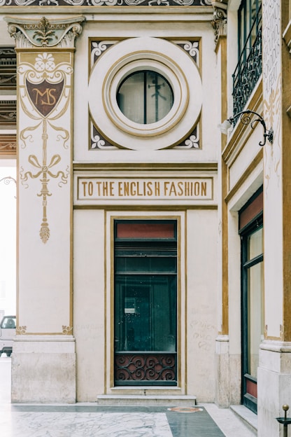 An ornate architectural facade featuring elegant designs and gold accents. A prominent circular window and a sign reading 'To the English Fashion' are focal points. Intricate detailing and embellishments are present, contributing to an elegant and vintage aesthetic.