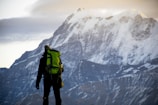 A person stands in front of a massive snow-capped mountain. The individual is wearing outdoor gear, including a bright green backpack, and appears to be looking towards the mountain. The scene suggests a moment of pause or contemplation amidst the mountaineering journey.