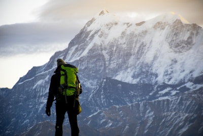 A person stands in front of a massive snow-capped mountain. The individual is wearing outdoor gear, including a bright green backpack, and appears to be looking towards the mountain. The scene suggests a moment of pause or contemplation amidst the mountaineering journey.
