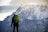 A person stands in front of a massive snow-capped mountain. The individual is wearing outdoor gear, including a bright green backpack, and appears to be looking towards the mountain. The scene suggests a moment of pause or contemplation amidst the mountaineering journey.