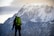 A person stands in front of a massive snow-capped mountain. The individual is wearing outdoor gear, including a bright green backpack, and appears to be looking towards the mountain. The scene suggests a moment of pause or contemplation amidst the mountaineering journey.