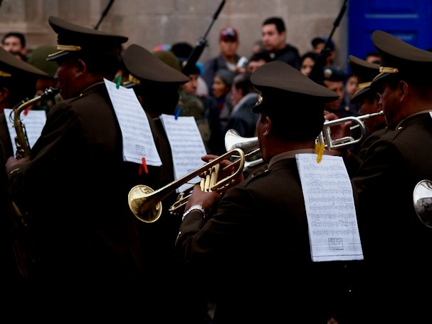 A group of uniformed musicians playing brass instruments in a parade or public performance. The musicians have sheets of music clipped to their backs, and they are closely surrounded by an audience. The setting appears to be an outdoor venue, possibly a historical or formal event.
