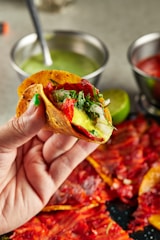 A hand is holding a vibrant taco filled with a variety of fresh ingredients including red chili peppers, onions, and green herbs. The background features two metal bowls containing green and red sauce, along with a lime placed nearby.