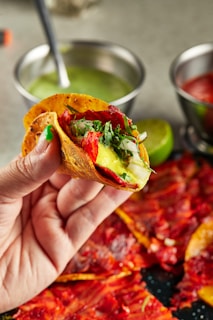 A hand is holding a vibrant taco filled with a variety of fresh ingredients including red chili peppers, onions, and green herbs. The background features two metal bowls containing green and red sauce, along with a lime placed nearby.