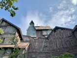 A large, old, wooden building complex with a sign that reads 'Mystery Mine 1892' on one of the structures. The buildings have a weathered and rustic appearance with some greenery in the foreground. The sky above is mostly clear with some clouds.