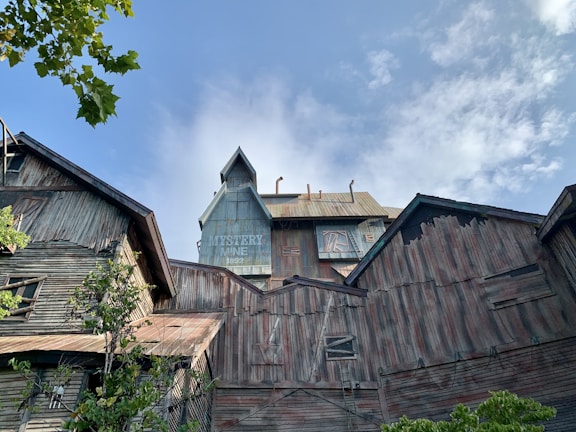 A large, old, wooden building complex with a sign that reads 'Mystery Mine 1892' on one of the structures. The buildings have a weathered and rustic appearance with some greenery in the foreground. The sky above is mostly clear with some clouds.