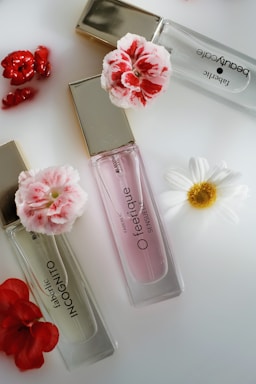 Three perfume bottles with elegant gold caps are arranged alongside vibrant red and white flowers on a sleek, white surface.