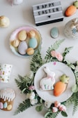An arrangement of Easter-themed items on a white surface, including decorated cookies shaped like a bunny and a carrot placed on a white plate surrounded by green leaves and pink flowers. There are pastel-colored eggs on a dish, a cup with colorful polka dots, a mini carrot decoration, and a lit-up sign with the text 'HAPPY EASTER' and a heart symbol.