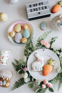 An arrangement of Easter-themed items on a white surface, including decorated cookies shaped like a bunny and a carrot placed on a white plate surrounded by green leaves and pink flowers. There are pastel-colored eggs on a dish, a cup with colorful polka dots, a mini carrot decoration, and a lit-up sign with the text 'HAPPY EASTER' and a heart symbol.